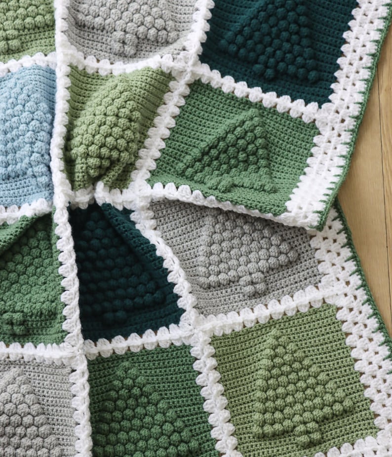 May include: A crocheted blanket with a white border and a geometric pattern of green, blue, and grey squares. The blanket is made with a textured stitch that creates a raised, bumpy surface.