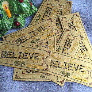May include: A collection of gold-colored Polar Express Believe Trip tickets. The text "Polar Express Believe Trip" is displayed on each ticket in a decorative font, with the word "BELIEVE" spelled out in black dots. A ceramic Christmas tree is in the background.