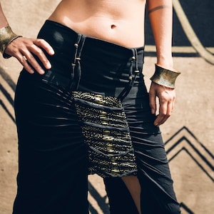 Op de afbeelding: Zwarte crop top en rok set met gouden accenten. De top heeft een uniek ontwerp en patroon, terwijl de rok een gedrapeerd ontwerp heeft met een bijpassend goudkleurig paneel. Goudkleurige manchetten maken de look compleet.