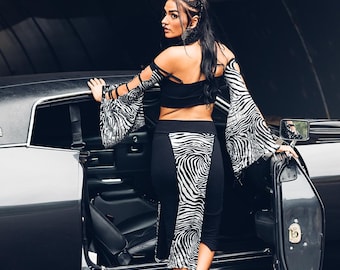 Op de afbeelding: Een zwarte crop top en potloodrok met een zebraprint paneel. De mouwen van de top zijn lang en uitlopend, met een bijpassende zebraprint. De persoon draagt zwarte laarzen en staat naast een zwarte auto.