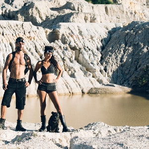 May include: A couple wearing black clothing and goggles stand in a white rocky landscape with a small body of water in the background. A small black dog stands between them.