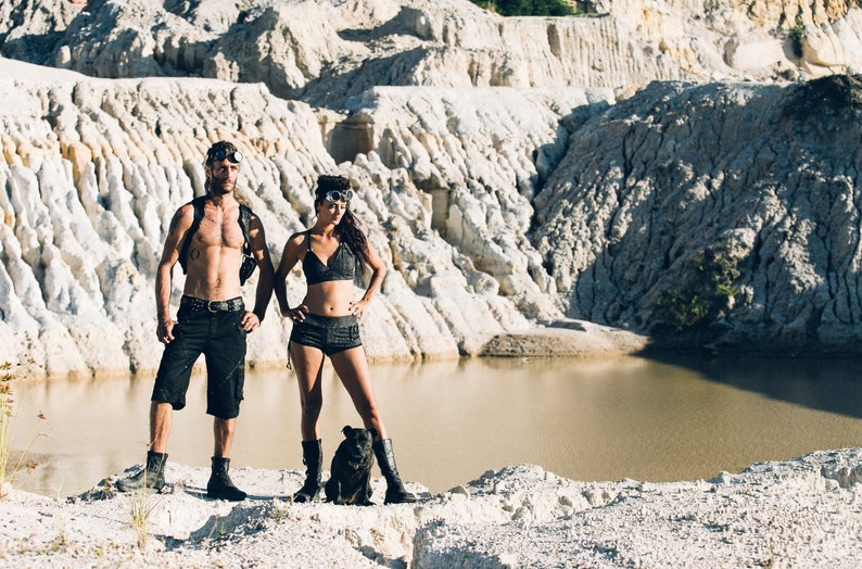 May include: A couple wearing black clothing and goggles stand in a white rocky landscape with a small body of water in the background. A small black dog stands between them.