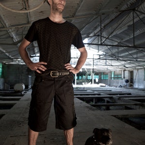 May include: A man wearing a black patterned shirt and black shorts stands in a large, industrial space. He is looking to the right of the frame. A black dog is sitting in front of him.