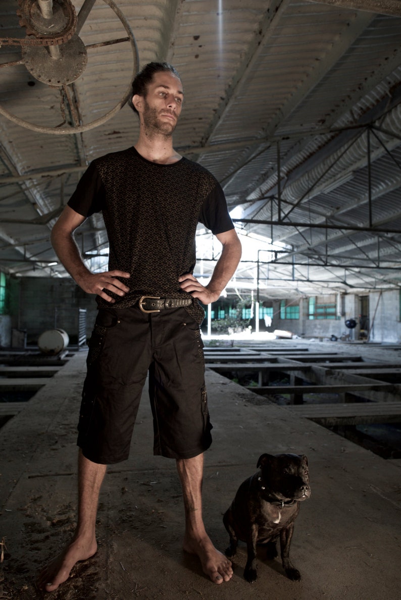 May include: A man wearing a black patterned shirt and black shorts stands in a large, industrial space. He is looking to the right of the frame. A black dog is sitting in front of him.
