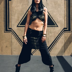 Op de afbeelding: Een vrouw draagt een zwarte crop top en bijpassende broek met wijde pijpen en gouden details. Ze draagt gouden armbanden en zwarte enkellaarsjes. De achtergrond is een betonnen muur met een zwart geometrisch ontwerp.