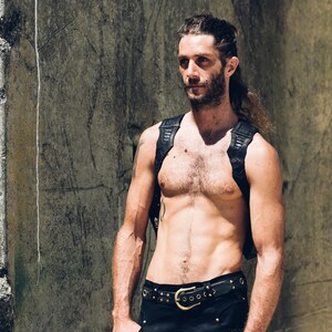 May include: A shirtless man with long hair and a beard is wearing a black leather harness and a black belt with a large silver buckle. He is standing in front of a concrete wall.