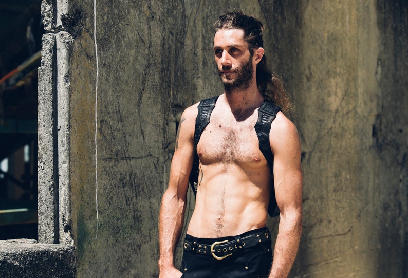 May include: A shirtless man with long hair and a beard is wearing a black leather harness and a black belt with a large silver buckle. He is standing in front of a concrete wall.
