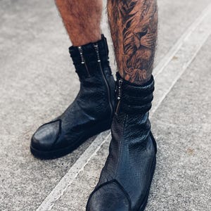 Pode incluir: Botas de couro preto com um padrão texturizado semelhante a pele de cobra. As botas têm fecho de correr e bico arredondado. A imagem mostra as botas dos tornozelos para baixo, com um fundo de concreto.