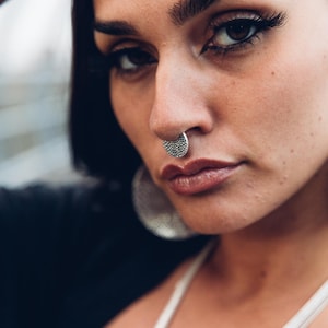 May include: Close-up of a person wearing a silver septum ring. The ring is a half-moon shape and is covered in small, round studs. The person has dark hair and is wearing a white top. The background is blurred.