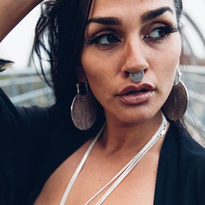 Może przedstawiać: Kobieta z ciemnymi włosami nosi srebrny kolczyk w nosie, kolczyki i naszyjnik. Kolczyk w nosie ma teksturowany, okrągły wzór. Kolczyki są duże, okrągłe i teksturowane. Naszyjnik ma wiele sznurków.