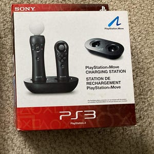 Op de afbeelding: Een PlayStation Move oplaadstation voor de PS3-console. De doos bevat een zwart oplaadstation met twee controllers en een apart oplaadstation. Het PlayStation Move-logo en het PS3-logo zijn zichtbaar.
