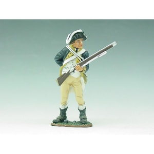 Puede incluir: Figura en miniatura de un soldado con uniforme histórico, sosteniendo un rifle. La figura viste una chaqueta azul oscuro, pantalones color crema y un sombrero tricornio. La figura se encuentra sobre una pequeña base verde.