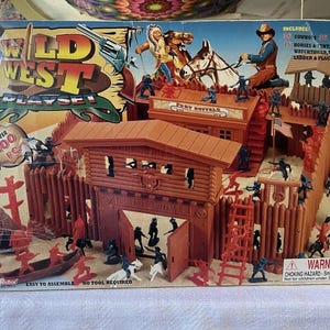 May include: A vintage Wild West playset box featuring a fort, cowboys, Native Americans, horses, and a canoe. The box includes the text "Wild West Playset" and "Over 100 pcs". The playset includes a fort, watchtower, totems, ladder, and flags.