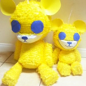 May include: Two yellow teddy bear piñatas with blue eyes and white faces. The larger bear is sitting next to a smaller one. Both have yellow ears and are made of a textured material. They are on a tiled floor.