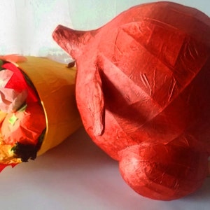 May include: A red paper mache pig with a tail made of twine, next to a yellow paper mache burrito filled with colorful paper mache fruit.