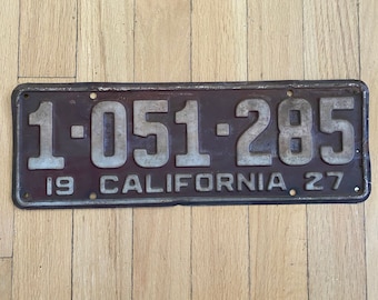May include: A vintage California license plate from 1927 with the number 1-051-285. The plate is made of metal and has a dark brown background with white lettering.