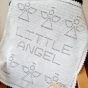 Peut inclure: Une couverture de bébé en crochet blanc avec un motif d'étoiles et les mots "Little Angel" au centre. Un petit ours en peluche est assis sur la couverture.
