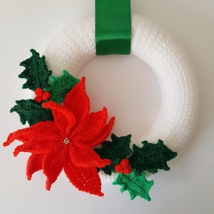 May include: A handmade Christmas wreath with a white base, adorned with a large red poinsettia and green holly leaves. A green satin ribbon is attached for hanging. The wreath is a festive holiday decoration.