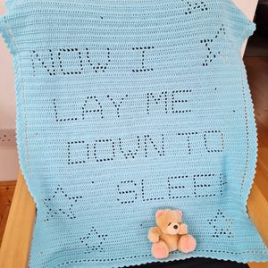 May include: A light blue crocheted baby blanket with the words "NOW I LAY ME DOWN TO SLEEP" stitched into the fabric. The blanket has star cutouts and a small teddy bear is resting on it.