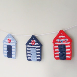 May include: A garland of crocheted beach huts in red, blue, and white with a white string connecting them. The huts have different details, including a life preserver, a door, and the word "Beach".