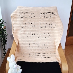Peut inclure: Une couverture en crochet crème, drapée sur une chaise en bois. La couverture affiche le texte "50% MOM 50% DAD" au-dessus de trois cœurs et les mots "100% PERFECT". Un lapin en peluche blanc est posé sur la chaise.