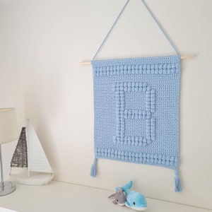 Puede incluir: Un tapiz de pared de crochet azul claro con un diseño texturizado y borlas. El tapiz está sujeto a una varilla de madera y tiene una cuerda para colgar.