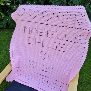 May include: A pink crocheted baby blanket with the text "Made with love on 2021" in black yarn.