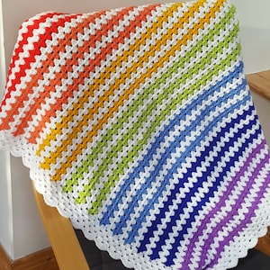 May include: A rainbow crochet blanket with a white border. The blanket is made with a granny square stitch and features a variety of colours, including red, orange, yellow, green, blue, indigo, and violet.