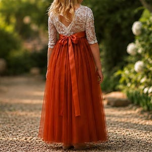 May include: A floor-length dress featuring a white lace bodice with three-quarter length sleeves and a deep V-back. The skirt is made of a solid, burnt orange tulle fabric, accented at the waist with a matching satin ribbon tied in a bow.