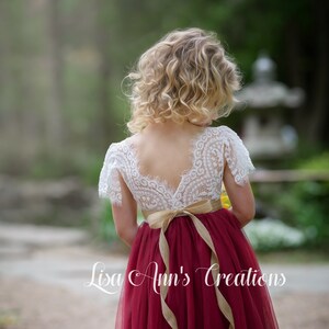 May include: A young girl wearing a white lace top with a burgundy tulle skirt and a brown sash. The skirt is long and flowing, and the sash is tied in a bow at the back. The girl is standing in a garden setting.