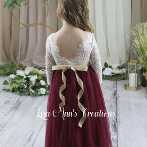 Flower girl dress in burgundy.  A young girl wearing a long-sleeved white lace dress with a burgundy tulle skirt. The dress has a V-shaped back and is tied with a tan ribbon at the waist.