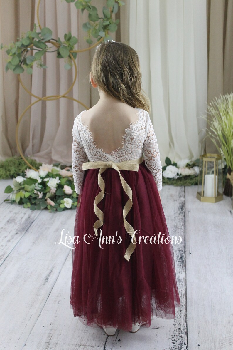 Flower girl dress in burgundy.  A young girl wearing a long-sleeved white lace dress with a burgundy tulle skirt. The dress has a V-shaped back and is tied with a tan ribbon at the waist.