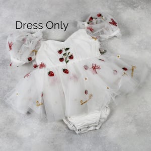 May include: White baby dress with strawberry embroidery and a matching bonnet. The dress features a tulle skirt and is adorned with red strawberries, bows, and the word "Sommer" in gold script. The text "Dress Only" is visible.