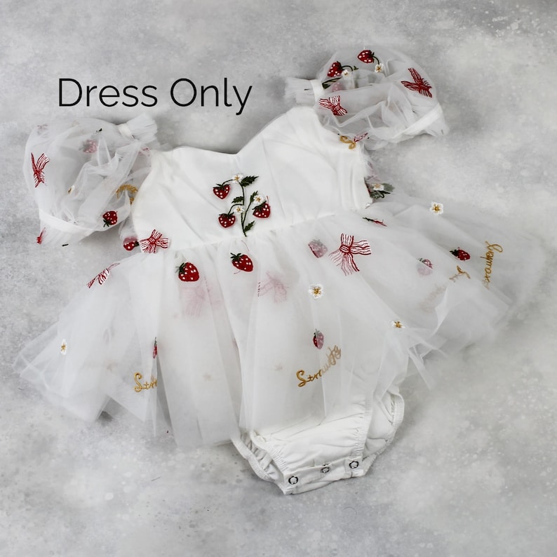 May include: White baby dress with strawberry embroidery and a matching bonnet. The dress features a tulle skirt and is adorned with red strawberries, bows, and the word "Sommer" in gold script. The text "Dress Only" is visible.