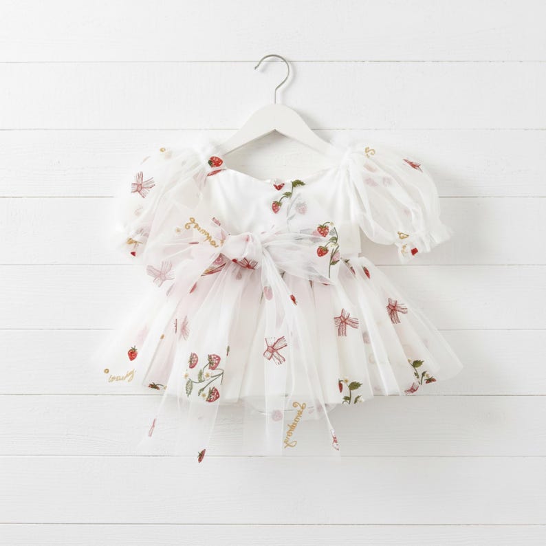 This enchanting strawberry birthday dress features exquisite, embroidered tulle adorned with strawberries, delicate bows, and a whimsical cake motif. The word "Strawberry" is beautifully embroidered throughout, creating a cohesive themed design perfect for first birthdays and milestone celebrations.