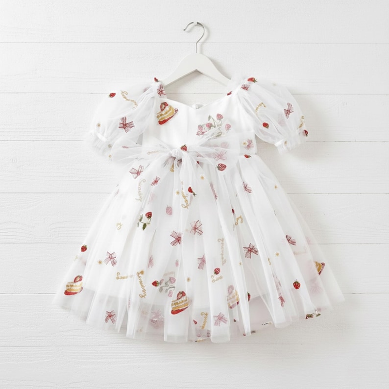 This enchanting strawberry birthday dress features exquisite, embroidered tulle adorned with strawberries, delicate bows, and a whimsical cake motif. The word "Strawberry" is beautifully embroidered throughout, creating a cohesive themed design perfect for first birthdays and milestone celebrations.