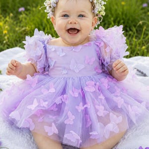 May include: A lavender dress featuring layered tulle fabric adorned with numerous three-dimensional butterfly appliqu&eacute;s, short puffed sleeves, and a round neckline. A floral crown with small white and purple blossoms rests on the head.