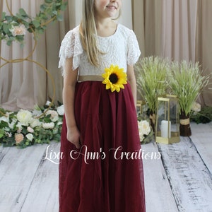 May include: A young girl wearing a white lace dress with a burgundy tulle skirt and a sunflower sash. The dress has short sleeves with lace ruffles. The girl is standing on a wooden floor with a floral backdrop.