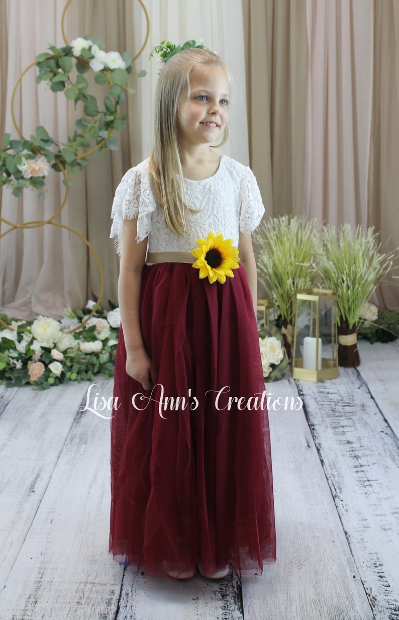 May include: A young girl wearing a white lace dress with a burgundy tulle skirt and a sunflower sash. The dress has short sleeves with lace ruffles. The girl is standing on a wooden floor with a floral backdrop.