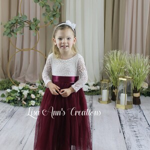 Burgundy flower girl dress.  A young girl wearing a white lace long-sleeved dress with a burgundy tulle skirt and a burgundy satin sash. The dress has a round neckline. The girl is wearing a white headband with a flower on top.