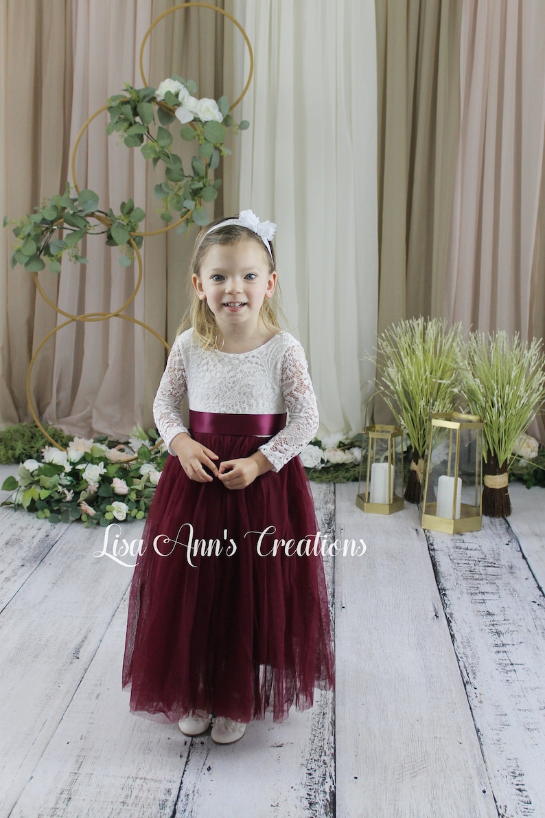 Burgundy flower girl dress.  A young girl wearing a white lace long-sleeved dress with a burgundy tulle skirt and a burgundy satin sash. The dress has a round neckline. The girl is wearing a white headband with a flower on top.