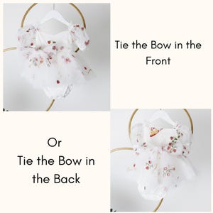 May include: White baby romper with floral embroidery, including strawberries and small flowers. The romper has puff sleeves and a tulle skirt. Text on the image reads "Tie the Bow in the Front" and "Tie the Bow in the Back".