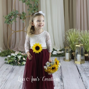 A young girl wearing a white lace long-sleeved dress with a burgundy tulle skirt and a sunflower sash. Dress for a flower girl in a wedding.