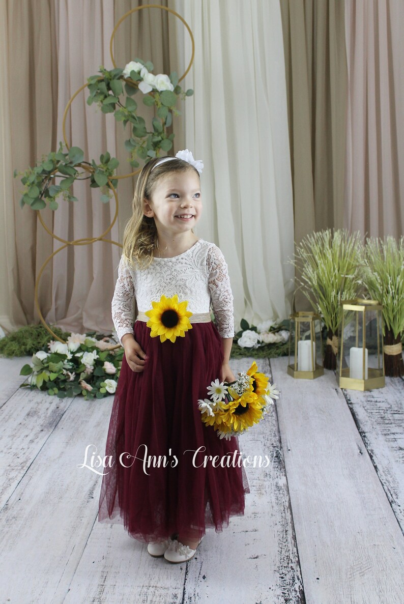 A young girl wearing a white lace long-sleeved dress with a burgundy tulle skirt and a sunflower sash. Dress for a flower girl in a wedding.