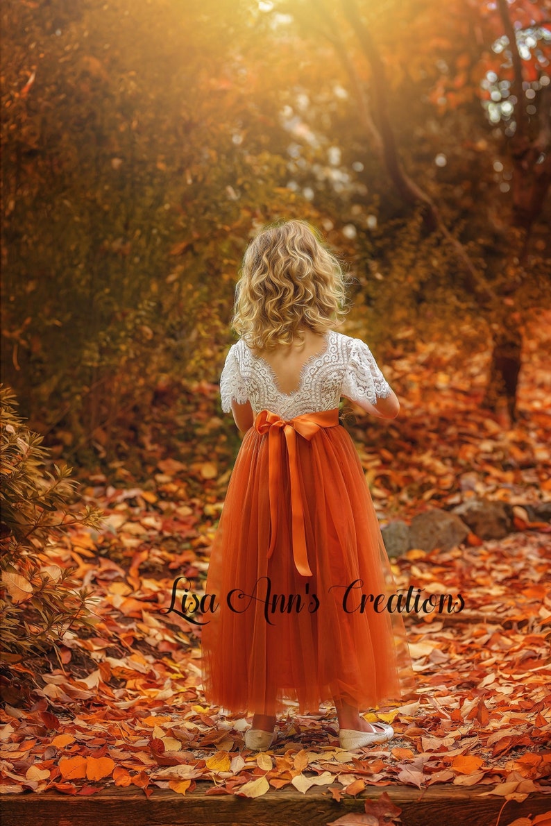 May include: A young girl in an orange tulle dress with a white lace top and a matching orange sash stands in a forest. The dress is knee-length. The background is filled with autumn leaves and foliage. The text "Lisa Ann's Creations" is at the bottom.