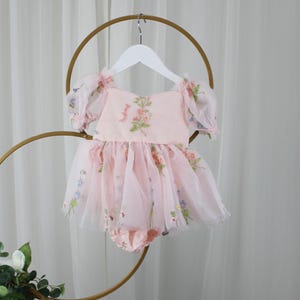 May include: A light pink baby romper with a floral design. The romper features a square neckline, short puff sleeves, and a tulle skirt. The tulle skirt is adorned with colorful floral accents. The romper is hanging on a white hanger.