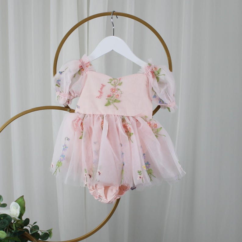 May include: A light pink baby romper with a floral design. The romper features a square neckline, short puff sleeves, and a tulle skirt. The tulle skirt is adorned with colorful floral accents. The romper is hanging on a white hanger.