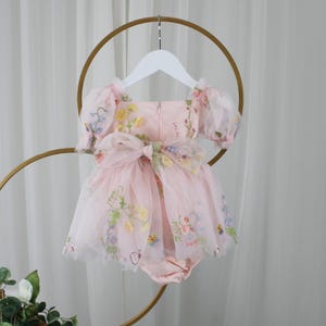 May include: A light pink baby romper with a floral design. The romper features a tulle overlay with embroidered flowers in yellow, blue, and purple. It has short, puffy sleeves and a large bow at the waist. The romper is hanging on a white hanger.