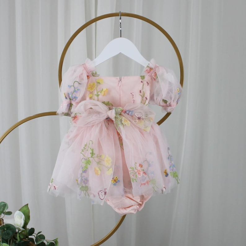 May include: A light pink baby romper with a floral design. The romper features a tulle overlay with embroidered flowers in yellow, blue, and purple. It has short, puffy sleeves and a large bow at the waist. The romper is hanging on a white hanger.