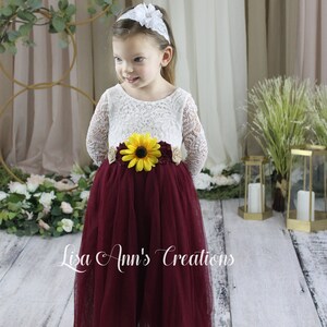Burgundy long sleeve dress for a flower girl. A young girl wearing a white lace long-sleeved dress with a burgundy tulle skirt. The dress has a sunflower and brown flower embellishment at the waist. The girl is wearing a white headband with a white flower on top.